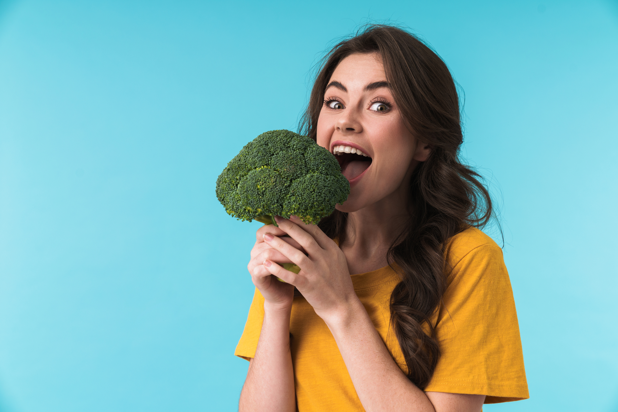 Image of optimistic positive young beautiful woman posing isolated over blue wall background holding broccoli.