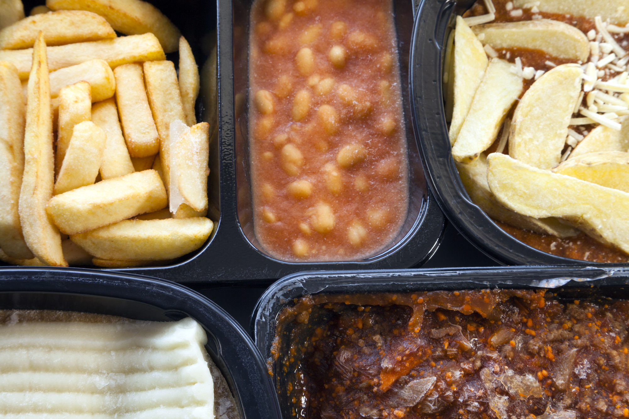 Selection of frozen, processed, ready made foods, consisting of potato chips, mince, lasagna in black, plastic containers . Microwave meals ready to heat up .