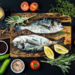 Fresh uncooked dorado or sea bream fish with lemon, herbs, oil, vegetables and spices on rustic wooden board over black backdrop