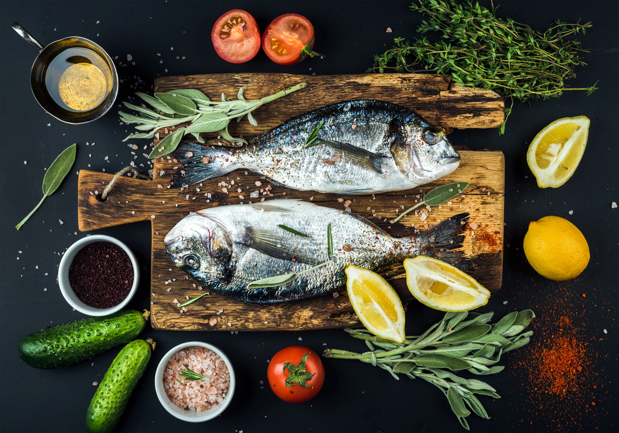 Fresh uncooked dorado or sea bream fish with lemon, herbs, oil, vegetables and spices on rustic wooden board over black backdrop