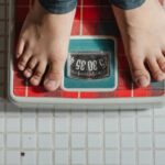 Weight Loss - From above crop anonymous barefoot child in jeans standing on weigh scales on tiled floor of bathroom