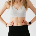 Navel - Faceless slim anonymous blond female in sports bra and black leggings in wearable bracelet showing perfect belly on white background while standing with hands on waist