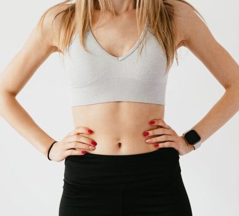 Navel - Faceless slim anonymous blond female in sports bra and black leggings in wearable bracelet showing perfect belly on white background while standing with hands on waist