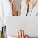 Blouses - Woman Looking on White Blouses Hanging on a Rack While Holding a Laptop