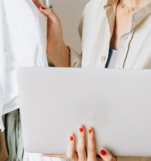 Blouses - Woman Looking on White Blouses Hanging on a Rack While Holding a Laptop