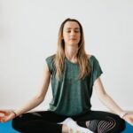 Mindfulness - Serene blond lady in sportswear sitting with crossed legs and closed eyes while practicing yoga at home near potted plants