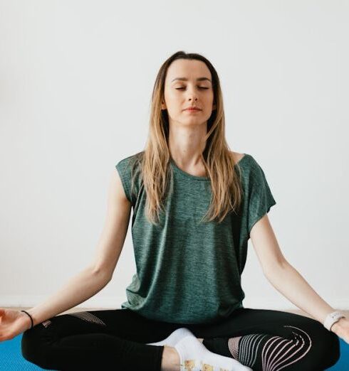 Mindfulness - Serene blond lady in sportswear sitting with crossed legs and closed eyes while practicing yoga at home near potted plants