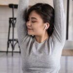 Mindfulness - Cheerful young ethnic female in earbuds sitting on yoga mat with crossed legs and closed eyes while listening to music and raising hands in apartment