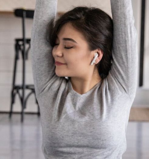 Mindfulness - Cheerful young ethnic female in earbuds sitting on yoga mat with crossed legs and closed eyes while listening to music and raising hands in apartment