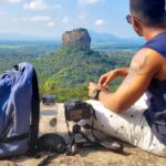 Traveling - Man Sitting on Top of Gray Cliff Mountain Beside Backpack, Water Bottle, and Camera