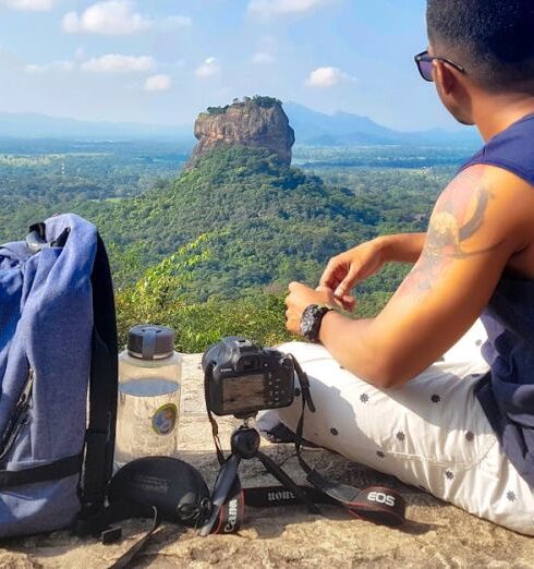 Traveling - Man Sitting on Top of Gray Cliff Mountain Beside Backpack, Water Bottle, and Camera