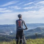 Cycling - Biker Holding Mountain Bike on Top of Mountain With Green Grass