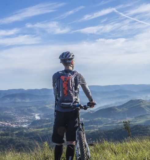 Cycling - Biker Holding Mountain Bike on Top of Mountain With Green Grass