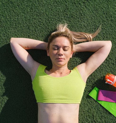 Training - Top view of peaceful female with closed eyes wearing sportive top lying with hands behind head while resting on sport ground near water bottles