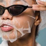 Routine - Self esteem young African American lady with moisturizing sheet mask and towel on hand wearing trendy sunglasses while standing against blue background in bathrobe after shower