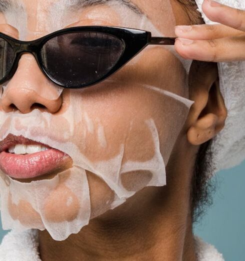 Routine - Self esteem young African American lady with moisturizing sheet mask and towel on hand wearing trendy sunglasses while standing against blue background in bathrobe after shower