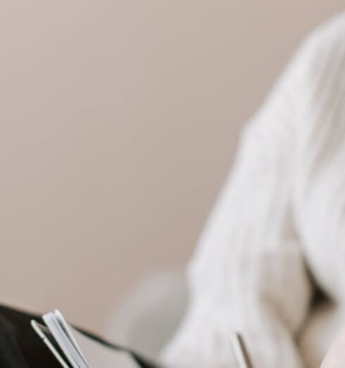 Journaling - Attentive female wearing eyeglasses and casual outfit sitting barefoot with crossed legs on comfortable couch in modern flat and taking notes in notebook with pen