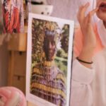 Self-Care - Young stylish female in eyeglasses and turban sitting on bed in bedroom and adjusting makeup while looking in mirror