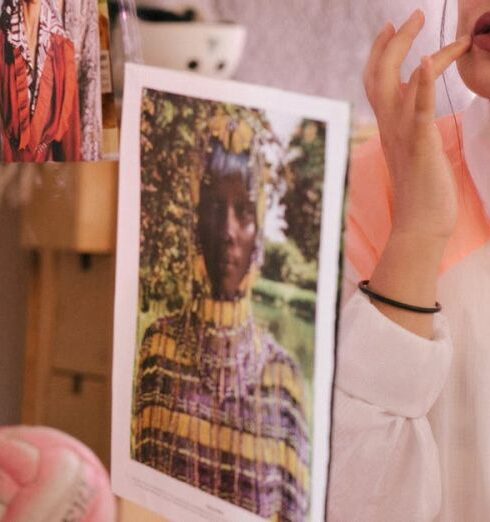 Self-Care - Young stylish female in eyeglasses and turban sitting on bed in bedroom and adjusting makeup while looking in mirror