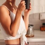 Routine - Side view of smiling lady in sleepwear standing in light kitchen while drinking cup of fresh beverage in morning near counter and cabinets