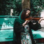 IRAs - Woman Playing Violin While Leaning On Green Vehicle