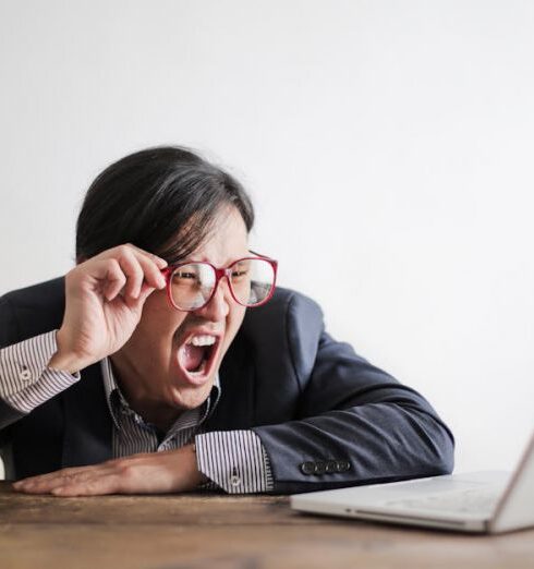 Mistakes - Modern Asian man in jacket and glasses looking at laptop and screaming with mouth wide opened on white background