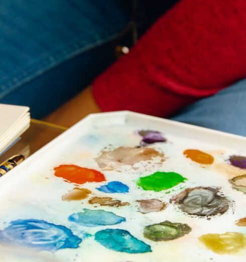 Watercolor - Crop anonymous painter sitting at table with bright paint palette near canvas while working on project