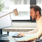 Emails - Man in White Dress Shirt Sitting on Black Rolling Chair While Facing Black Computer Set and Smiling