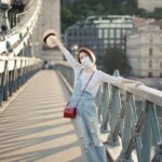 Excursions - Full body of anonymous happy young female traveler in stylish outfit and face mask standing on bridge crossing river with outstretched arm in ancient city