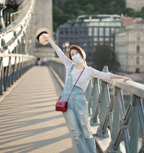 Excursions - Full body of anonymous happy young female traveler in stylish outfit and face mask standing on bridge crossing river with outstretched arm in ancient city