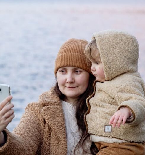 Family Cruises - Calm woman carrying adorable child on hands and taking selfie on smartphone near river and mountains