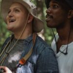 Surprise Trip - Side view astonished diverse males in casual summer clothes standing with photo camera in verdant forest and looking up in surprise