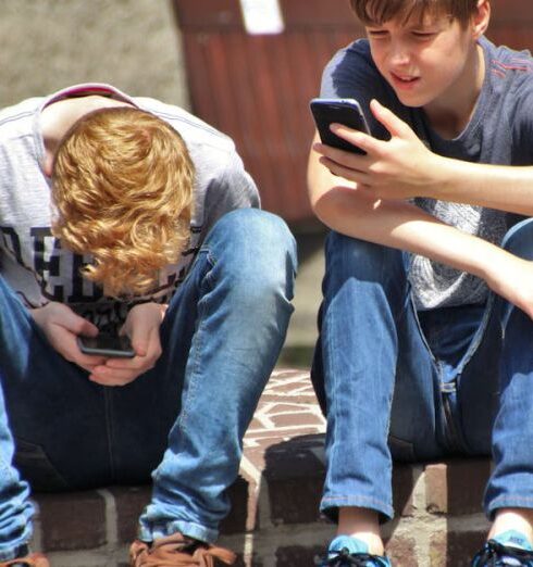 Teenagers - 2 Boy Sitting on Brown Floor While Using Their Smartphone Near Woman Siiting on Bench Using Smartphone during Daytime