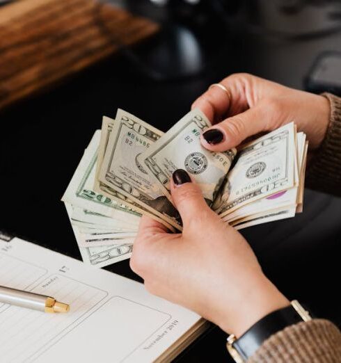 Budget - Unrecognizable elegant female in sweater counting dollar bills while sitting at wooden table with planner and pen