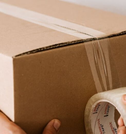 Packing - Crop anonymous male using tape to seal packed cardboard box against white wall while moving into new place