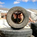 Campsites - Pensive boys looking through big black wheel of vehicle under vibrant blue sky with clouds in encampment