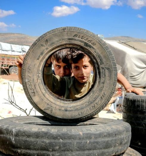 Campsites - Pensive boys looking through big black wheel of vehicle under vibrant blue sky with clouds in encampment