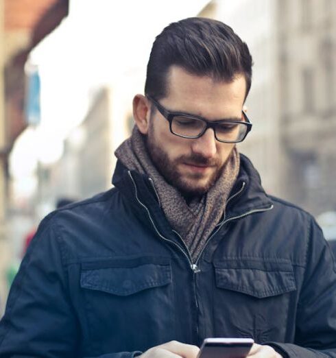 Jacket - Man Wearing Black Zip Jacket Holding Smartphone Surrounded by Grey Concrete Buildings