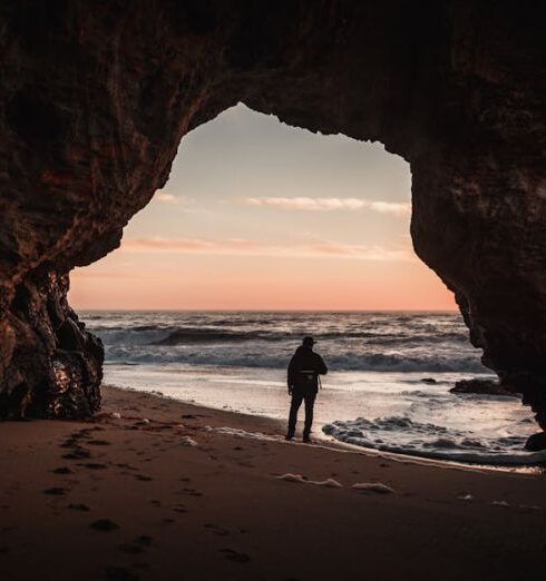 Hidden Gems - Person Standing at the Entrance of the Cave on Shore