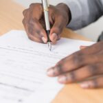 Document - Person Holding Gray Twist Pen and White Printer Paper on Brown Wooden Table