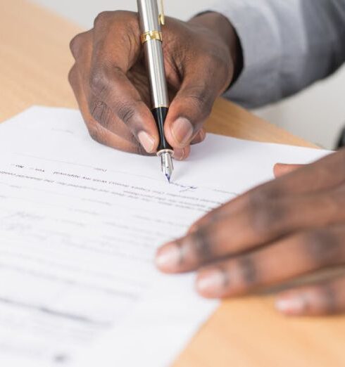 Document - Person Holding Gray Twist Pen and White Printer Paper on Brown Wooden Table