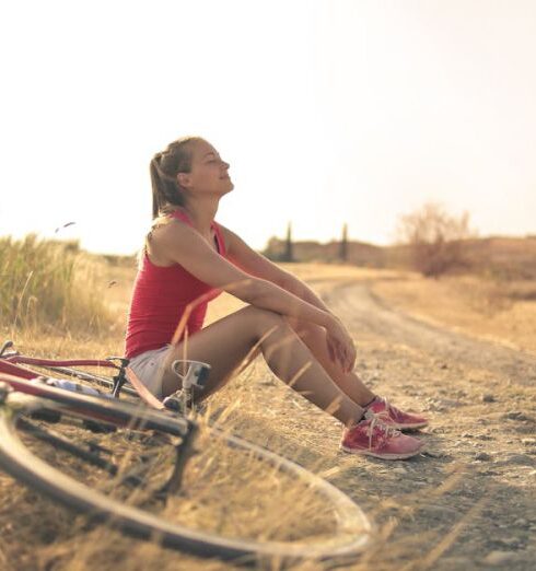 Wellness - Full body of female in shorts and top sitting on roadside in rural field with bicycle near and enjoying fresh air with eyes closed