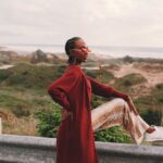 Sunglasses - Side view of young stylish African American female traveler in trendy outfit and sunglasses sitting on road metal fence during trip near ocean against cloudy sky