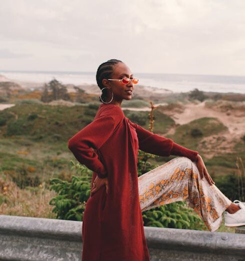 Sunglasses - Side view of young stylish African American female traveler in trendy outfit and sunglasses sitting on road metal fence during trip near ocean against cloudy sky