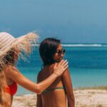 Bikinis - Woman in Sun Hat and Red Bikini Applying Lotion on a Woman's Back in Green Bikini