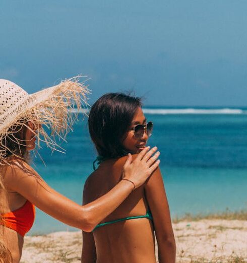 Bikinis - Woman in Sun Hat and Red Bikini Applying Lotion on a Woman's Back in Green Bikini