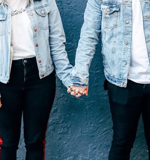 Denim - Two Person Standing and Holding Their Hands Beside Blue Wall