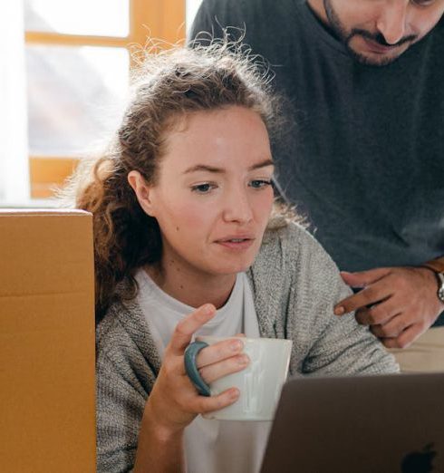 Black Friday - Thoughtful woman surfing internet on netbook while sitting with cup of hot drink near ethnic boyfriend in watch and casual wear standing near cardboard boxes after moving into new flat