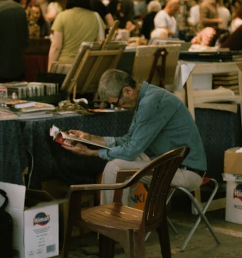 Book Fairs - Man Reading a Magazine Sitting at a Book Stall