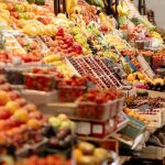 Grocery Stores - Woman in Red Clothes and Beret Walking Beside the Fruits Store